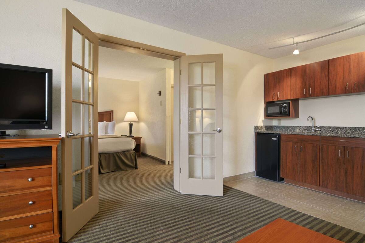 A hotel room with a bed, bedside table, lamp, TV, wooden dresser, and a kitchen area with a microwave and dishwasher.