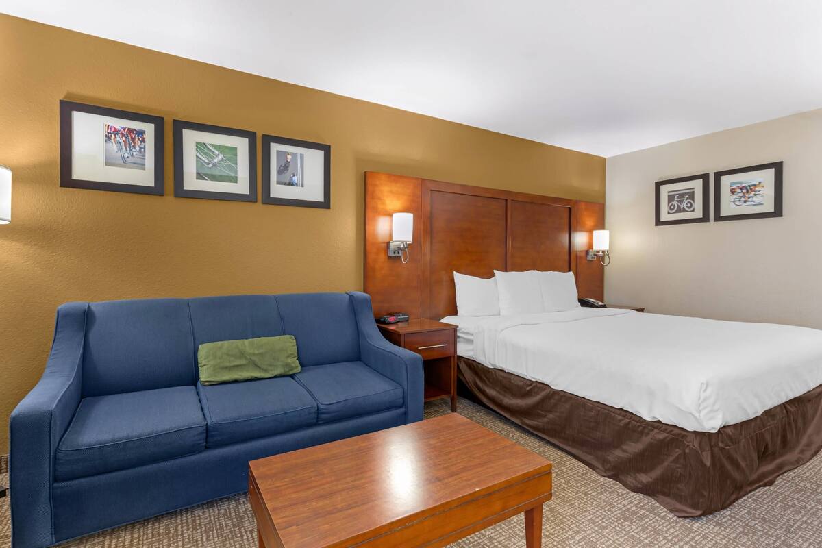 A hotel room with a blue sofa, a wooden coffee table, a bed with white bedding, and framed pictures on the wall.