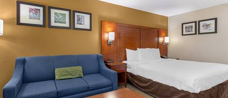 Habitación de hotel con un sofá azul, una mesa de centro de madera, una cama con ropa de cama blanca y cuadros en la pared.