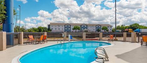 A swimming pool with lounge chairs, a fence, and a multi-story building in the background.
