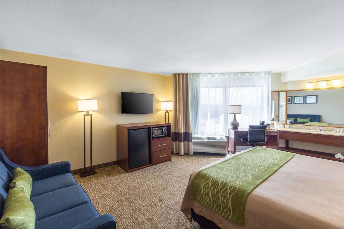 A hotel room with a bed, a desk, a television, a minibar, and a seating area with a sofa and armchair.