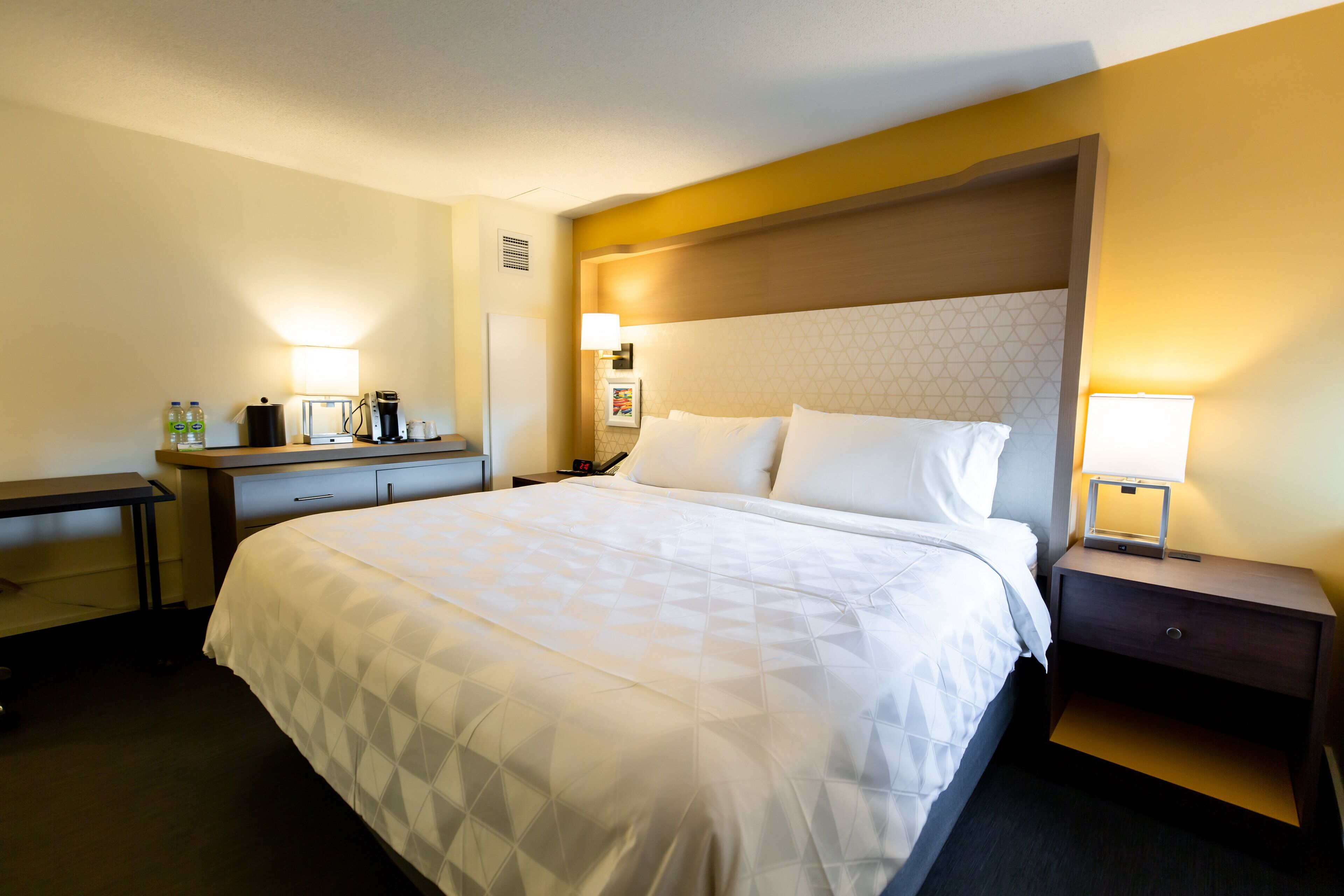 A hotel room with a large bed, a bedside table, a nightstand, a desk, and a lamp.