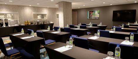 A conference room with multiple tables, chairs, and water bottles. There is a coffee station and a large screen on the wall.