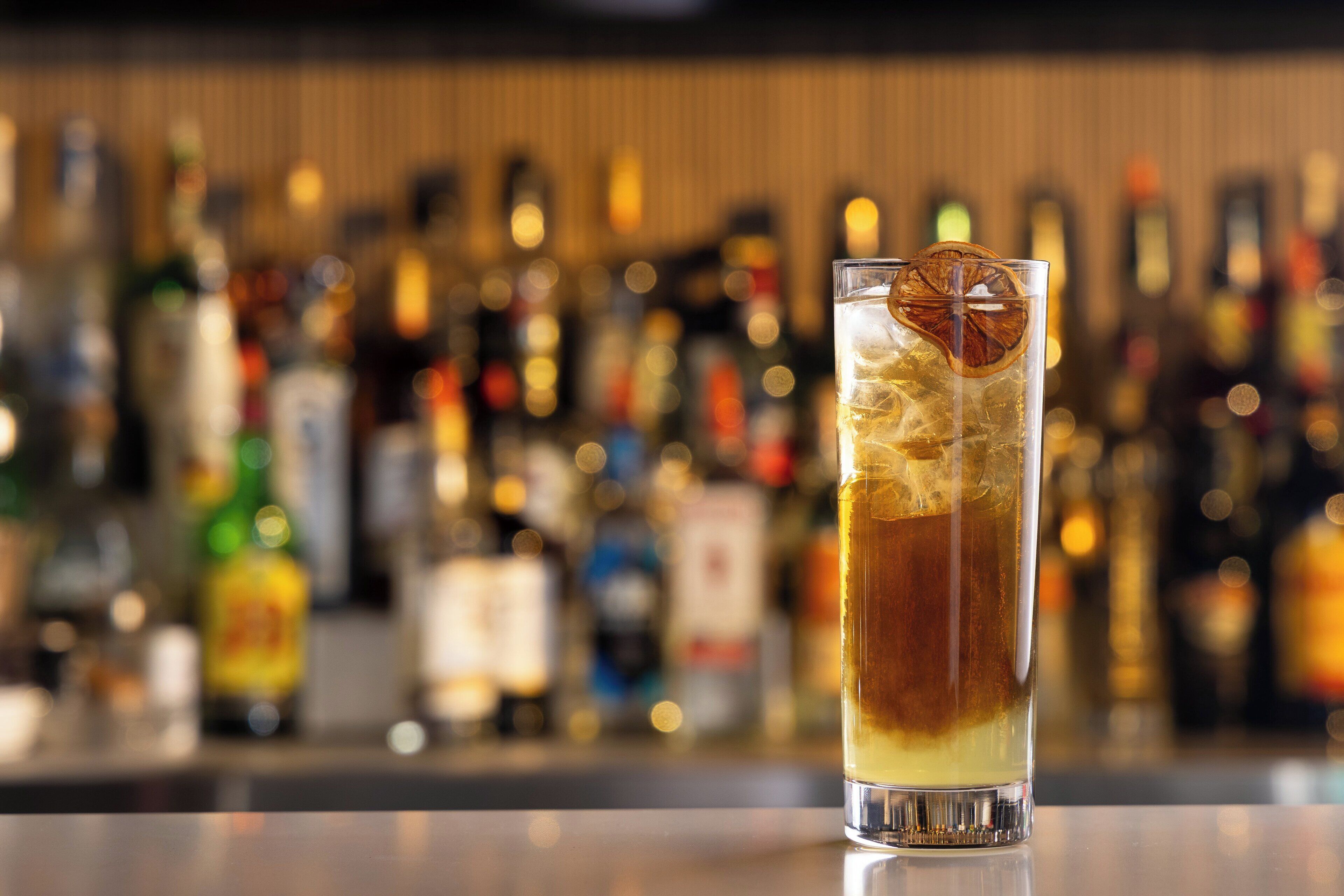 A bar setting with a tall glass of a layered drink, ice, and a dried orange slice garnish.