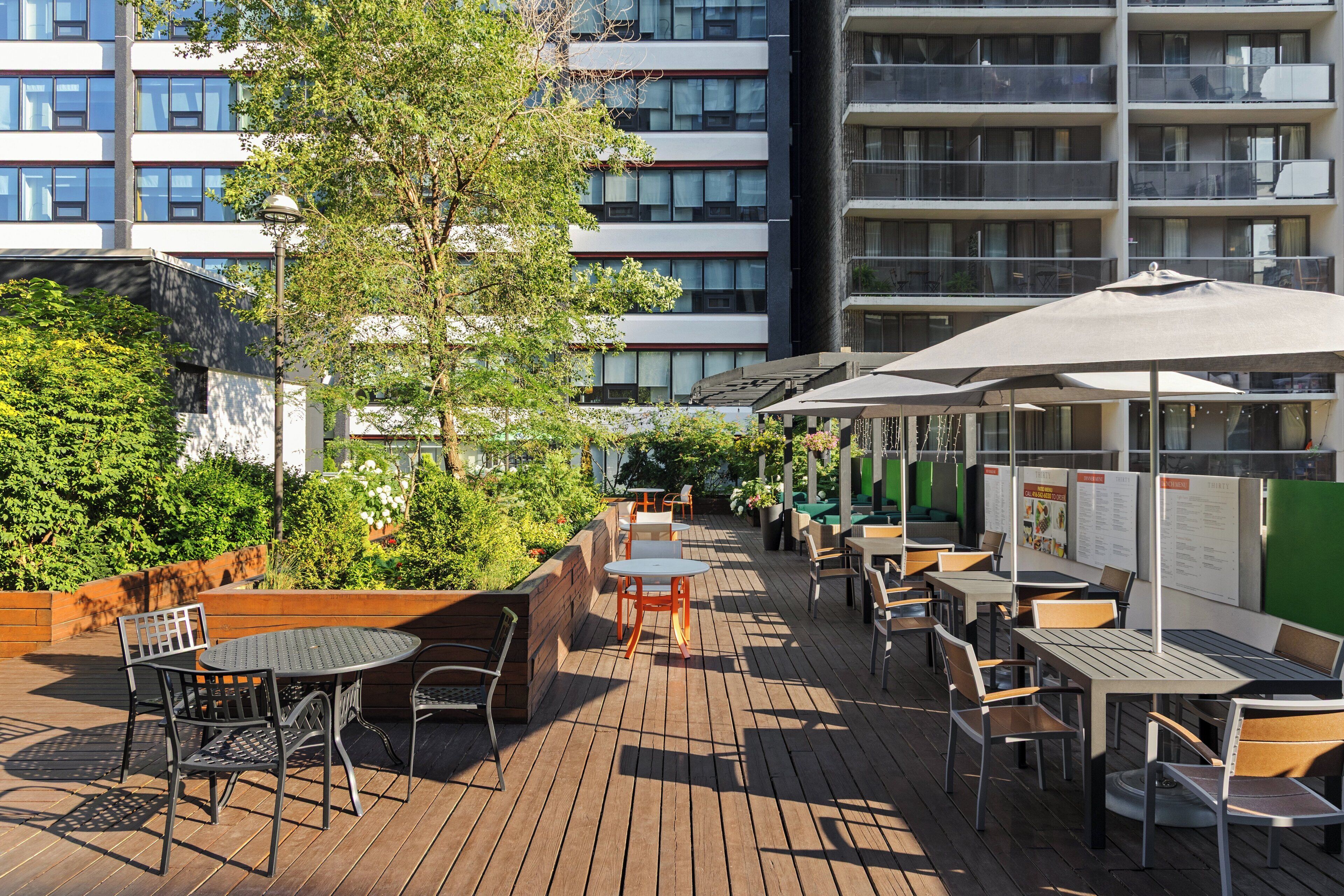 An outdoor patio area with tables and chairs, surrounded by greenery and a building in the background.