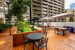 Een terras buiten met houten vloer, metalen tafels en stoelen, en groene planten.