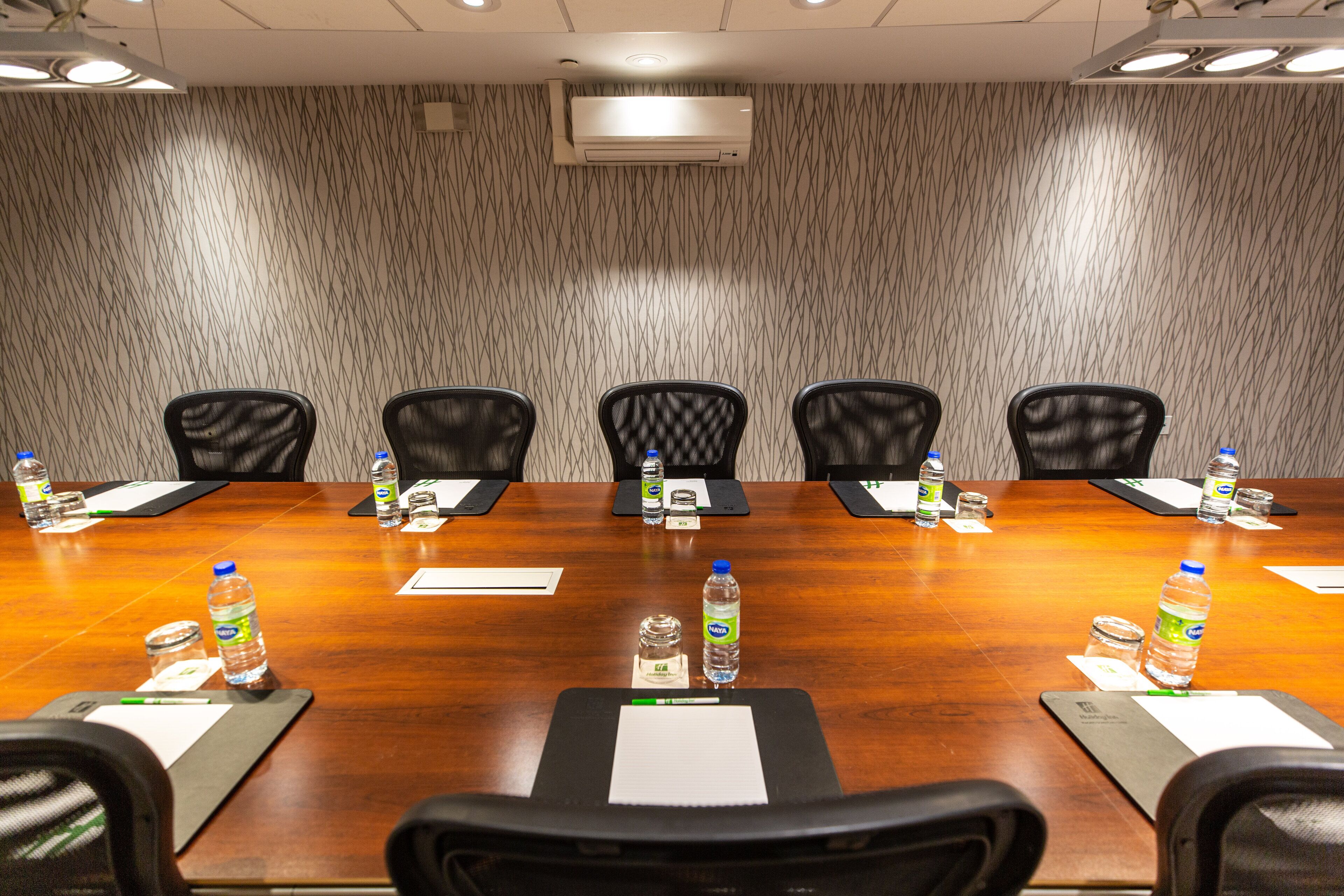 A conference room with a long wooden table, six chairs, water bottles, name tags, and a projector screen.