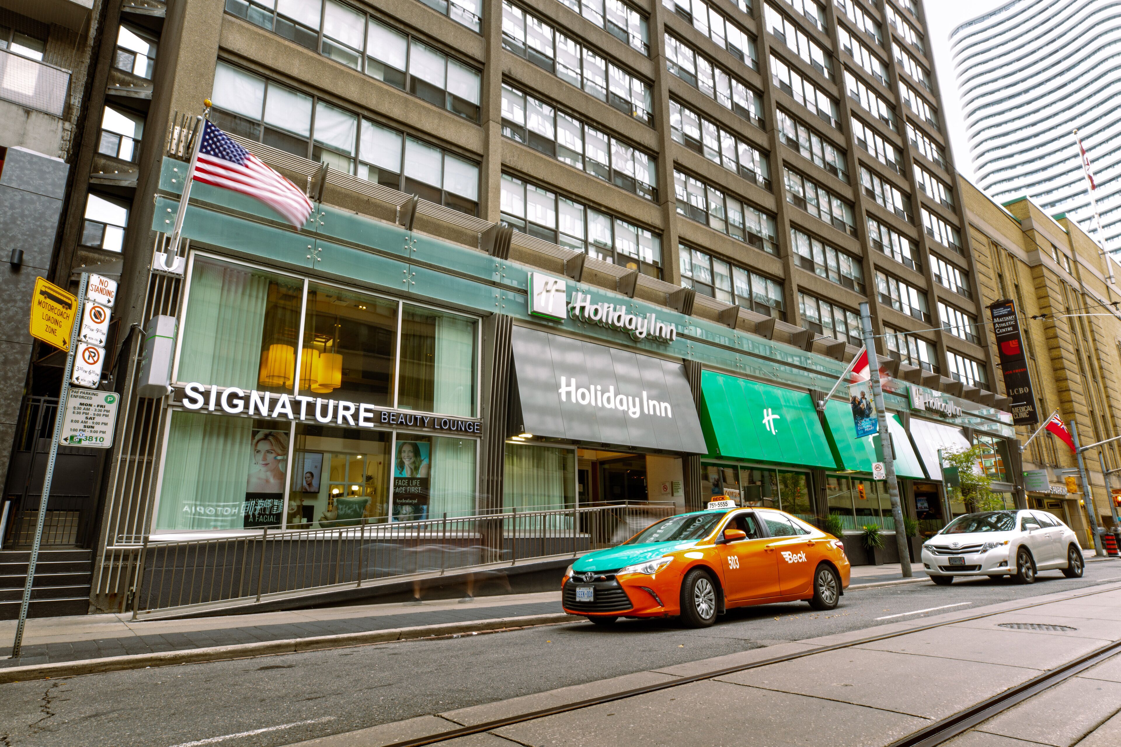 Un hotel Holiday Inn con un letrero que dice 'Signature Beauty Lounge', una bandera estadounidense y un taxi en la entrada.