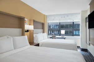 Quarto de hotel com duas camas, uma escrivaninha, uma cadeira e uma janela grande com vista para a cidade.