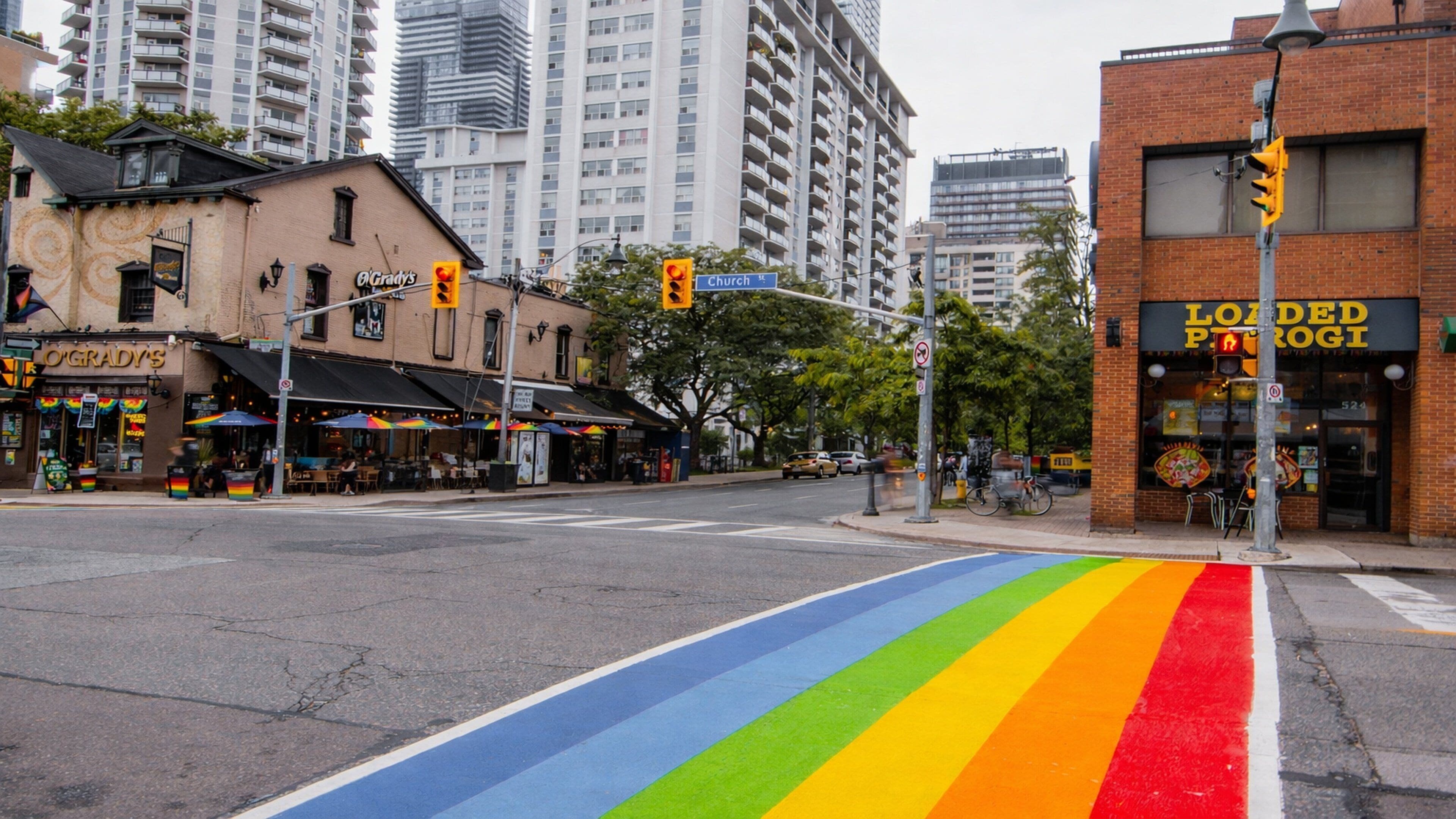 A street with a rainbow bike lane, a restaurant named 'O'Grady's', and a building with 'LOADED PIZZA' signage.