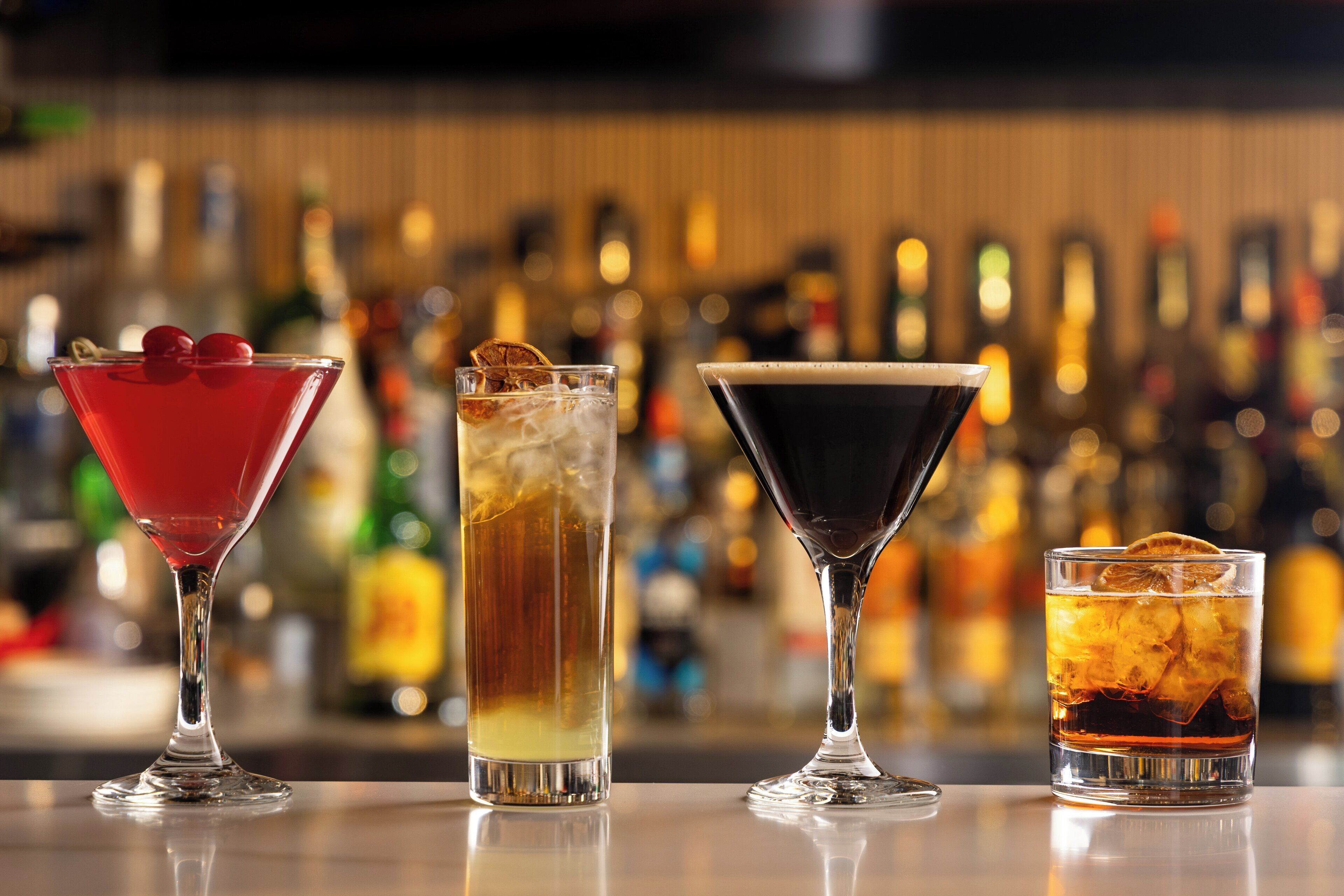 Four different cocktails on a bar counter with a blurred background of bottles.