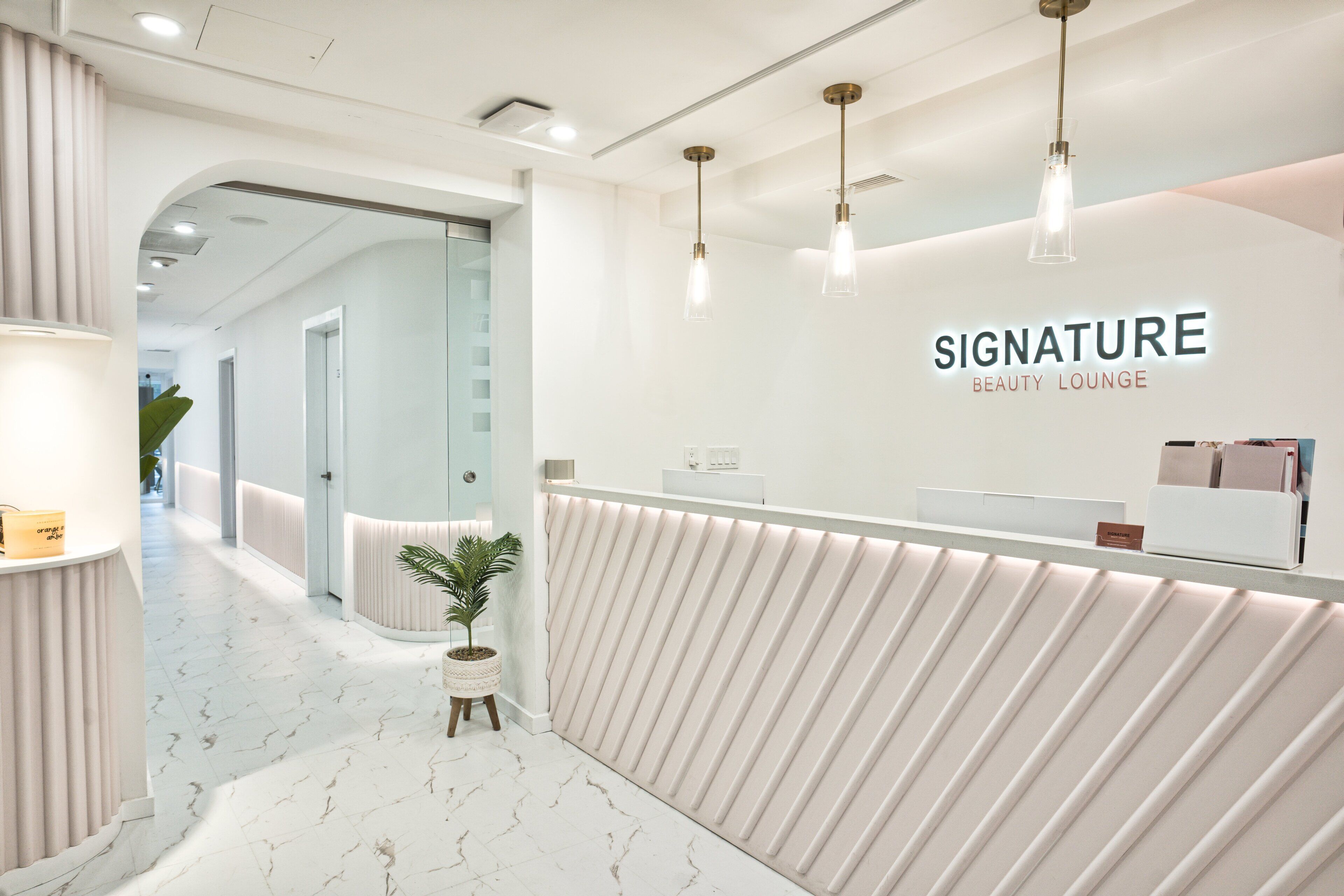 Un área de recepción de diseño moderno, con mostrador de recepción, planta en maceta y un cartel en la pared que dice "SIGNATURE BEAUTY LOUNGE".