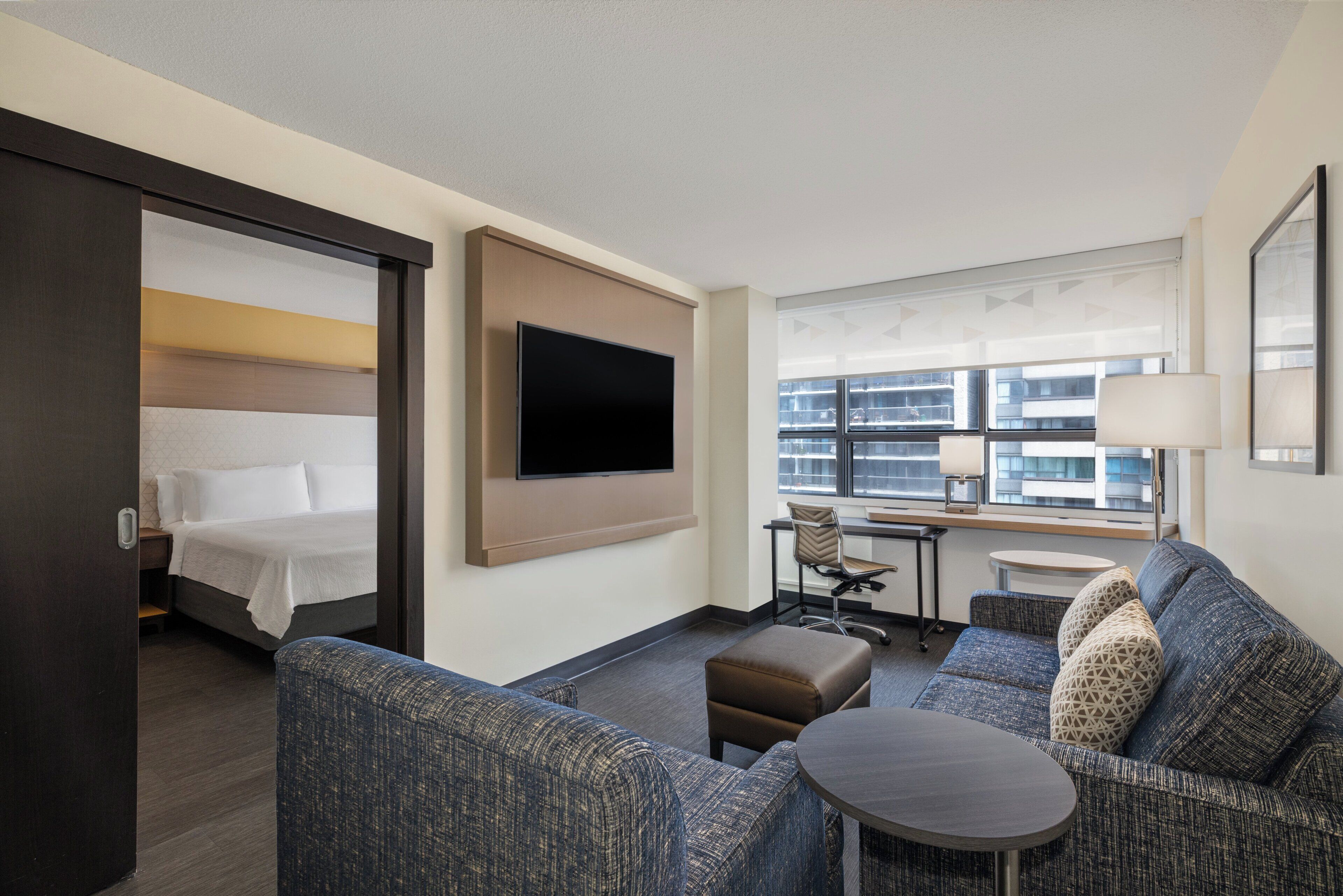 A hotel room with a bed, a sofa, a small table, a desk, and a TV mounted on the wall.