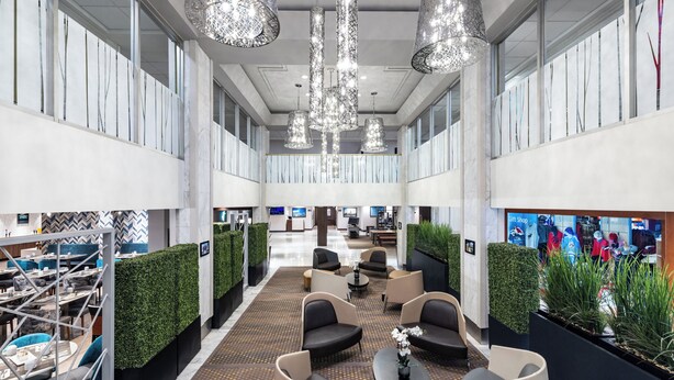 Un vestíbul d'hotel modern amb una zona de seients central, parets verdes i una cafeteria al fons.