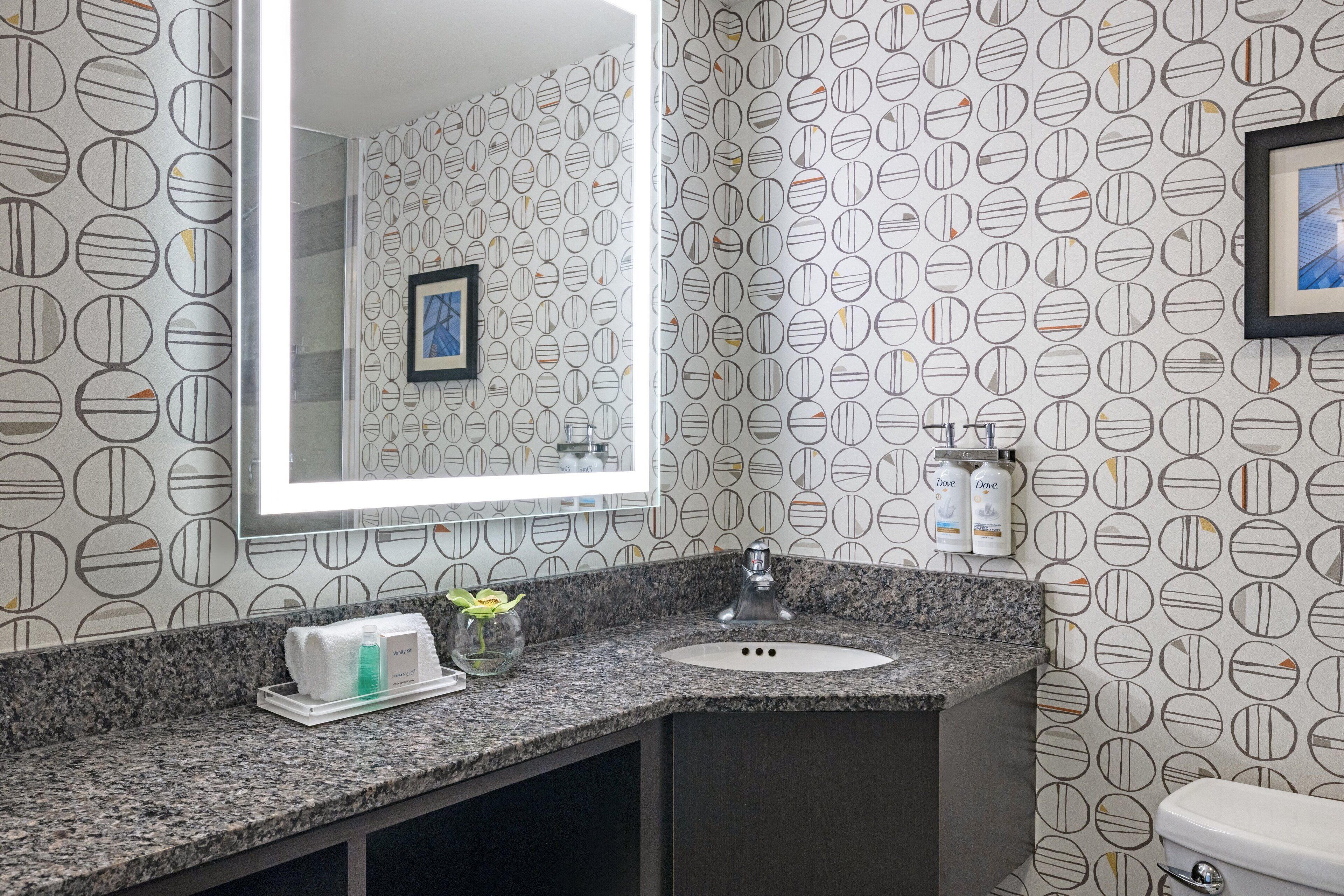 Un baño con encimera de granito, lavabo, espejo y papel tapiz estampado.