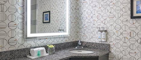 Un baño con encimera de granito, lavabo, espejo y papel tapiz estampado.