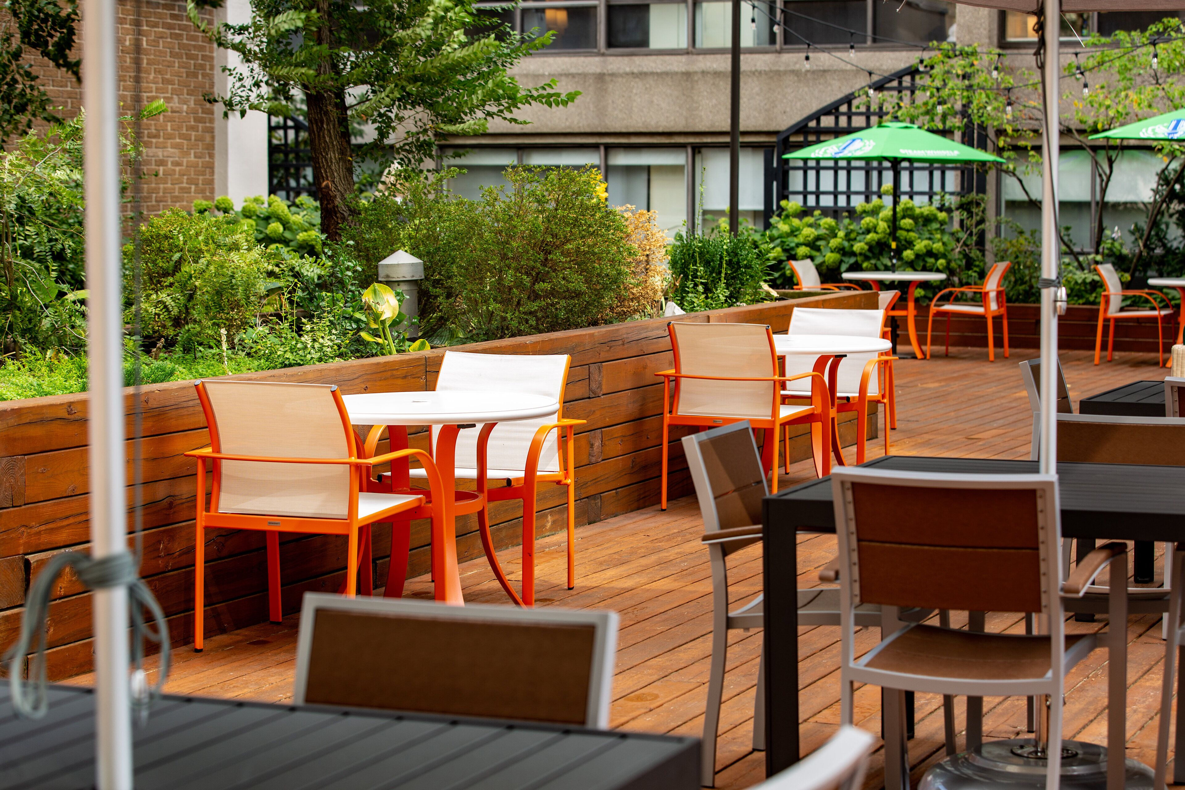 An outdoor patio with wooden tables and chairs, surrounded by greenery and a brick wall.