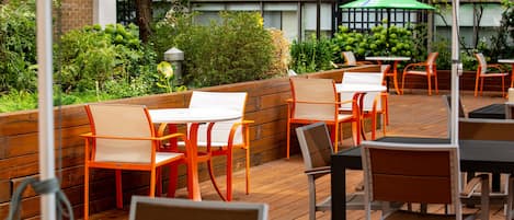 An outdoor patio with wooden tables and chairs, surrounded by greenery and a brick wall.