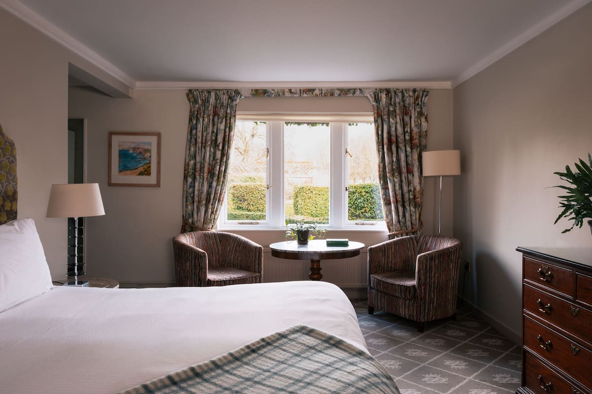 A hotel room with a bed, two armchairs, a small table, a lamp, a dresser, and a window with curtains.