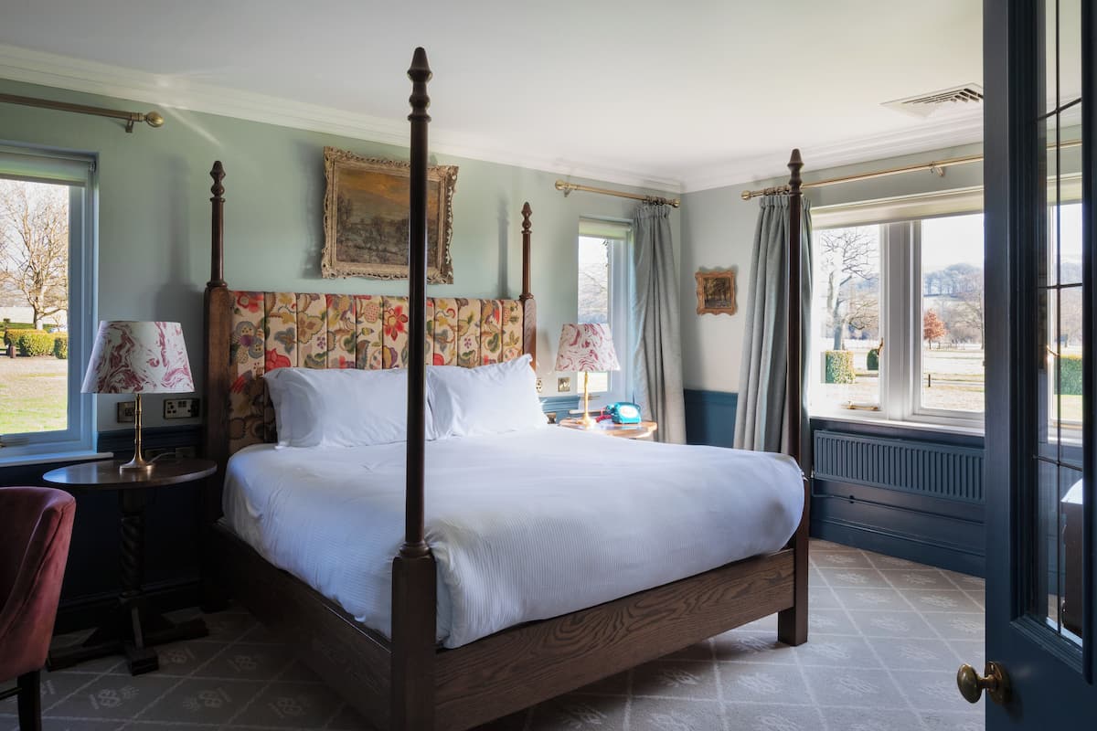 A four-poster bed with a floral headboard, a bedside table with a lamp, and a view of the outdoors through a large window.
