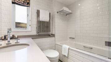 Un baño moderno con pared de azulejos blancos, cabina de ducha con mampara de vidrio, inodoro blanco y lavamanos curvo.