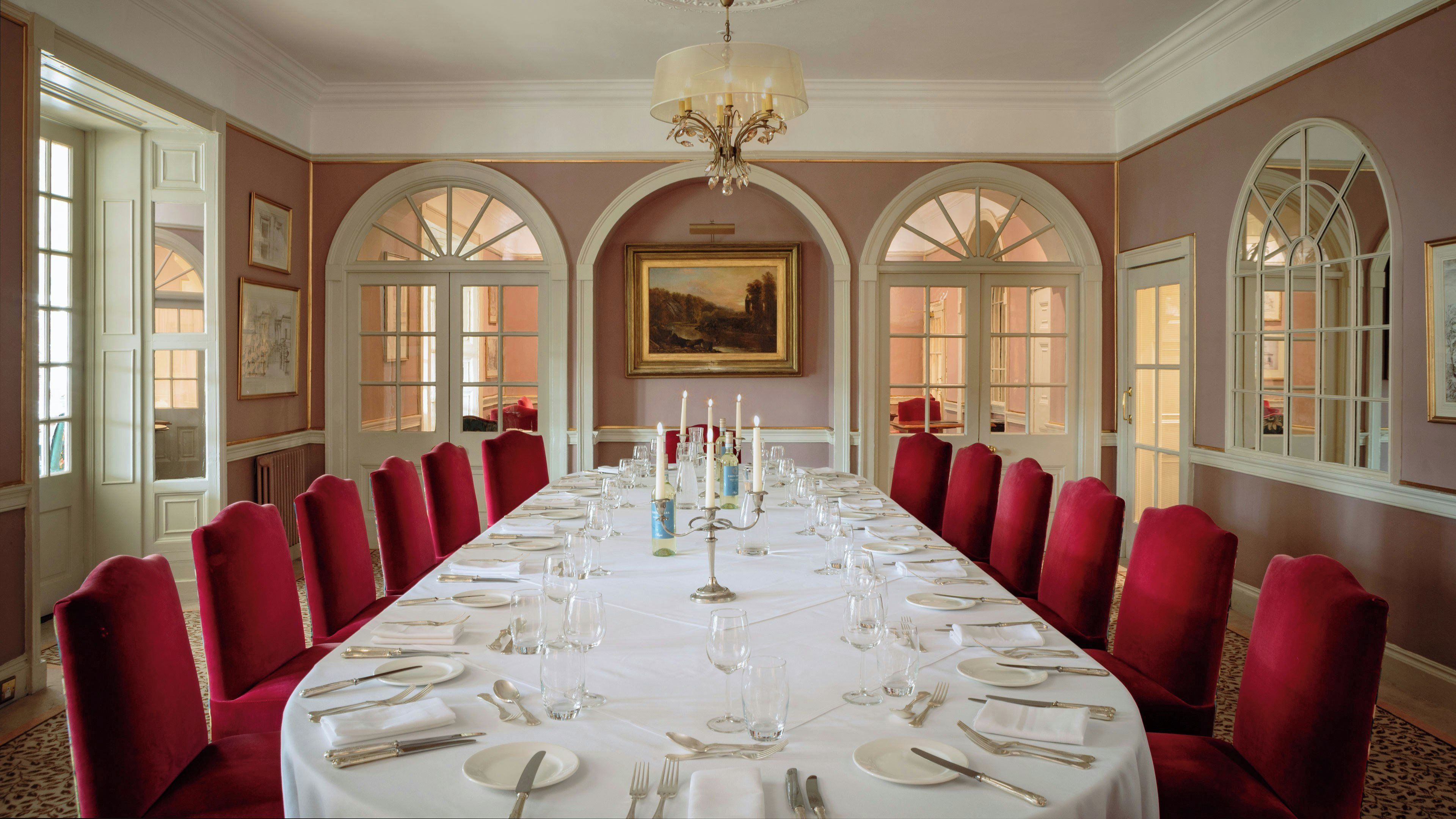 A formal dining room with a long table set for a meal, red upholstered chairs, and a chandelier.