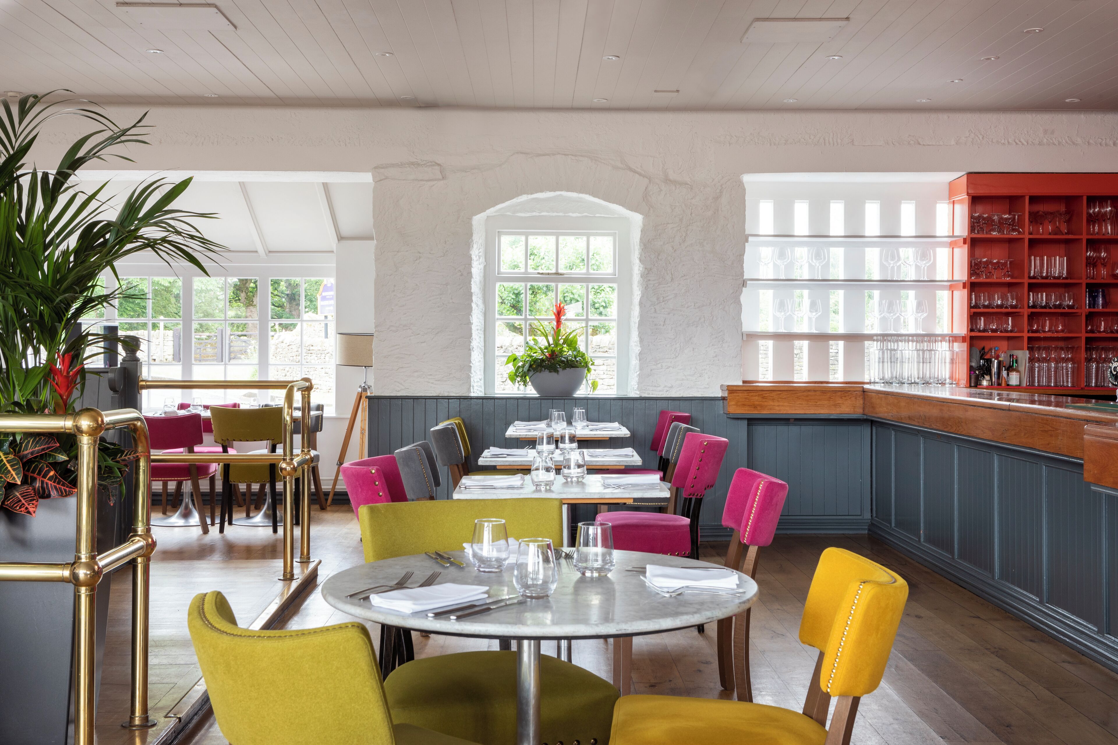 A restaurant with round tables, colorful chairs, and a bar area with shelves of glasses.