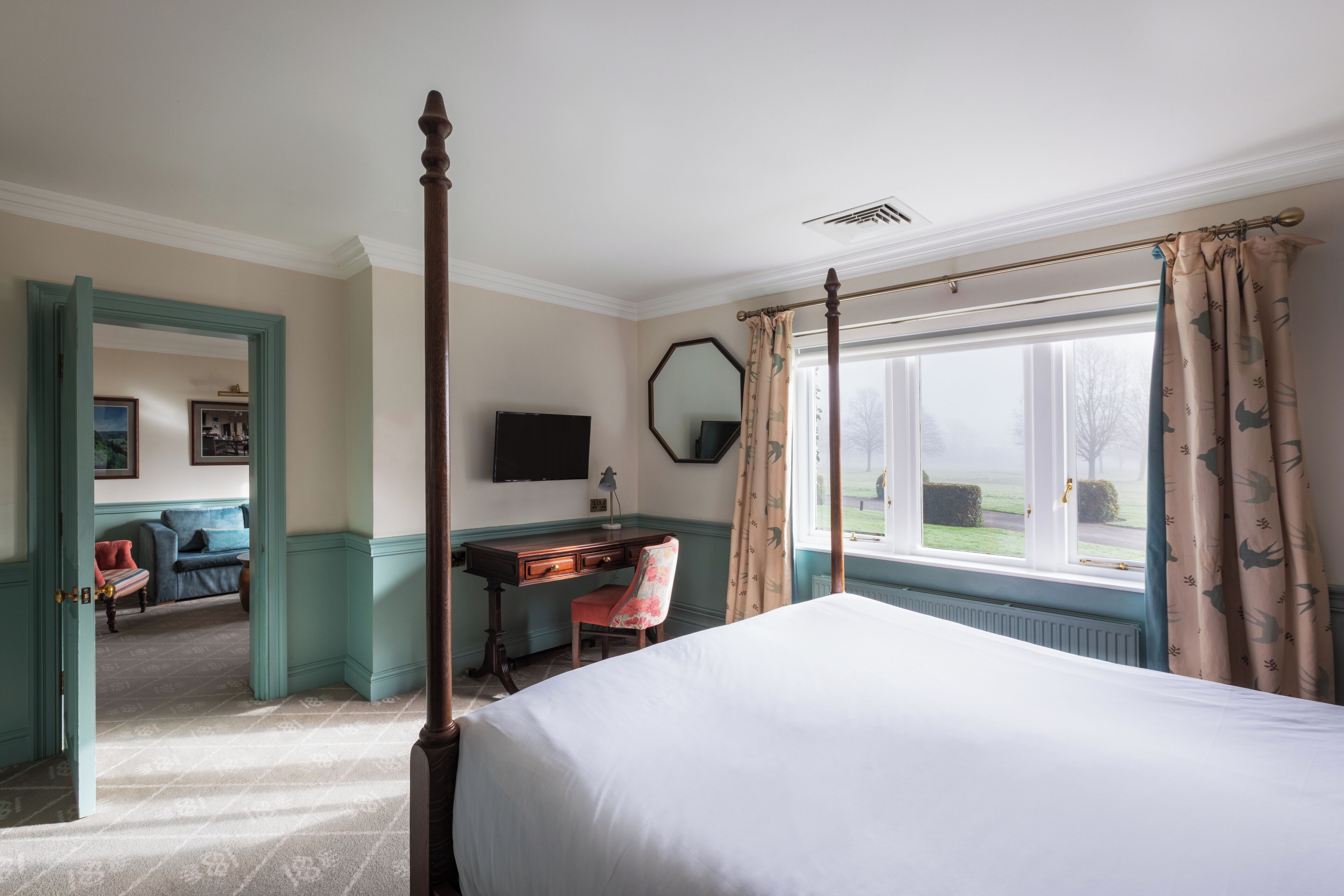 A bedroom with a four-poster bed, a desk with a chair, a television, and a large window with curtains.