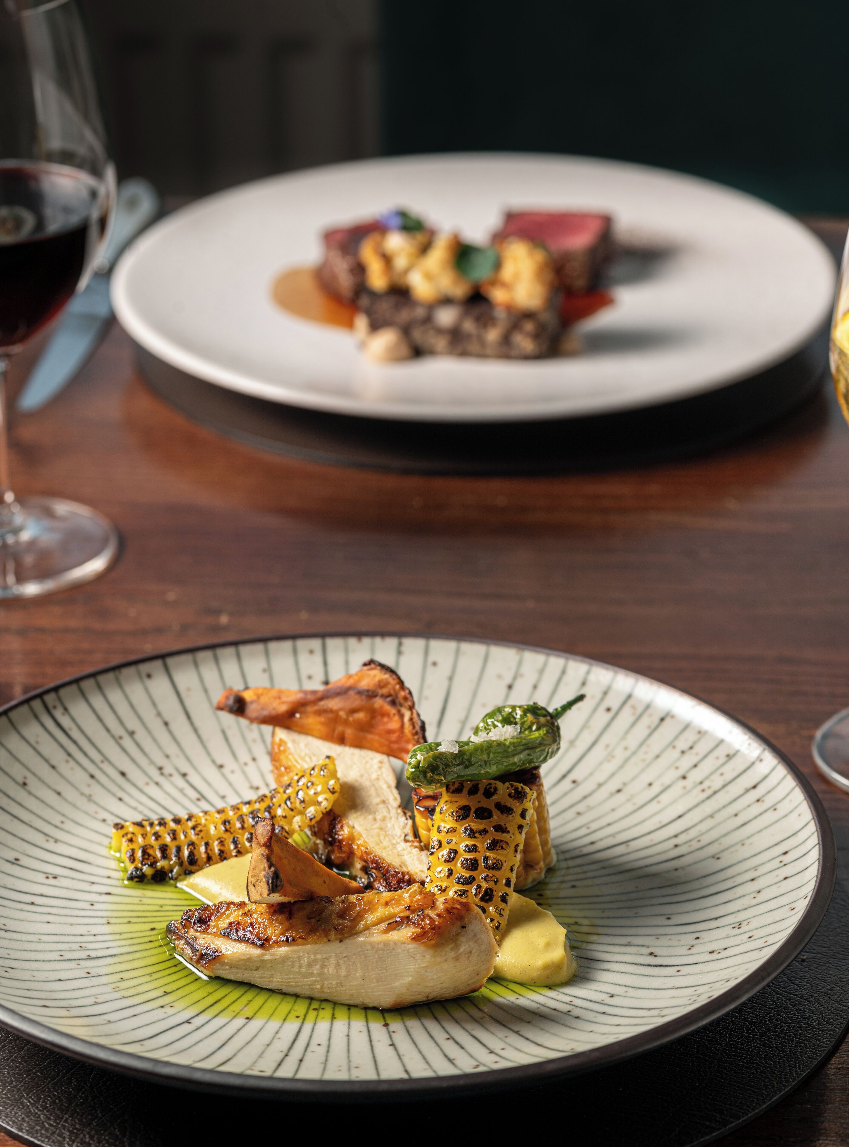 A dining table with two plates of food, one with grilled chicken and corn, and another with steak and vegetables.