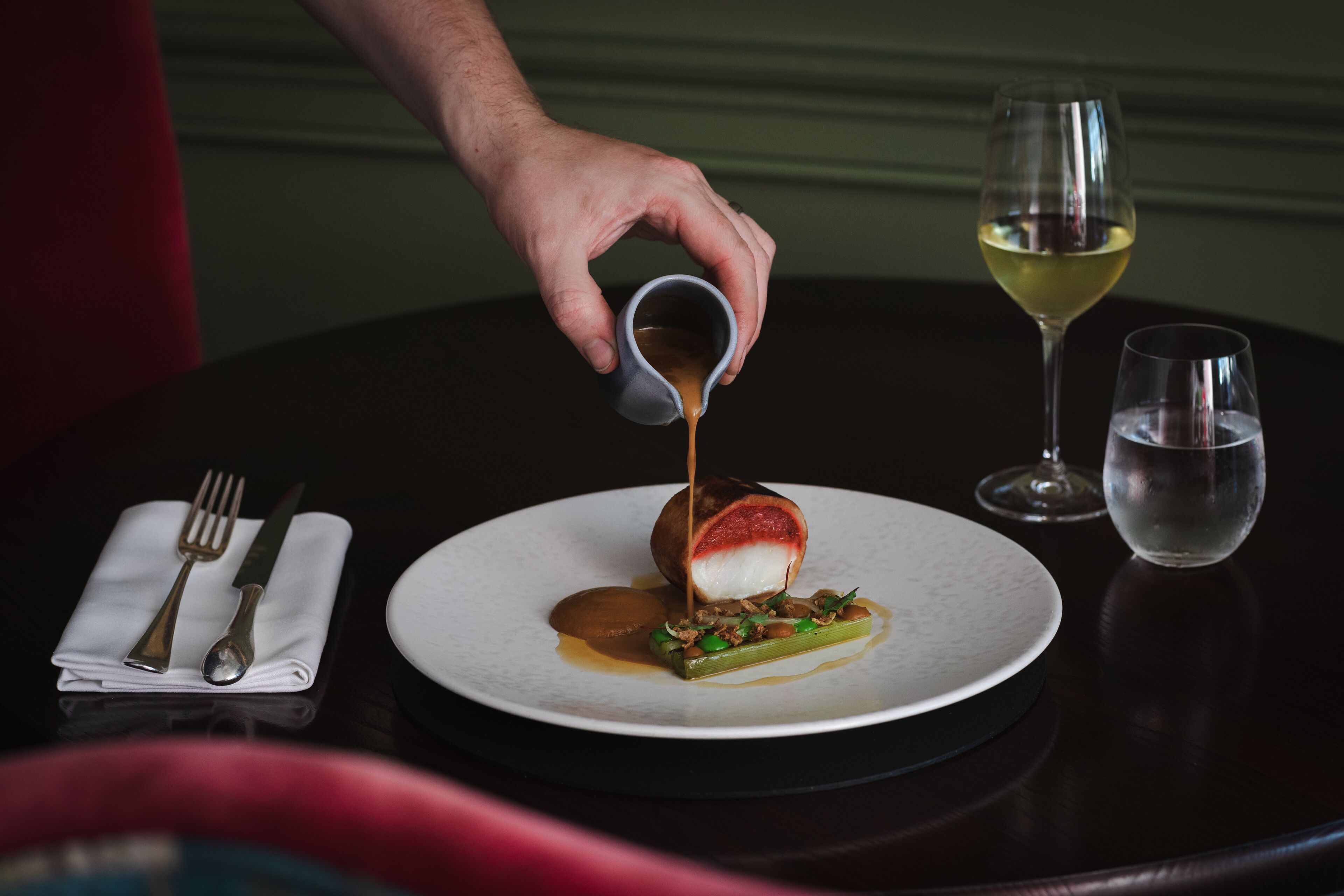 A dining table with a plate of food, a glass of wine, and a cup of sauce being poured.