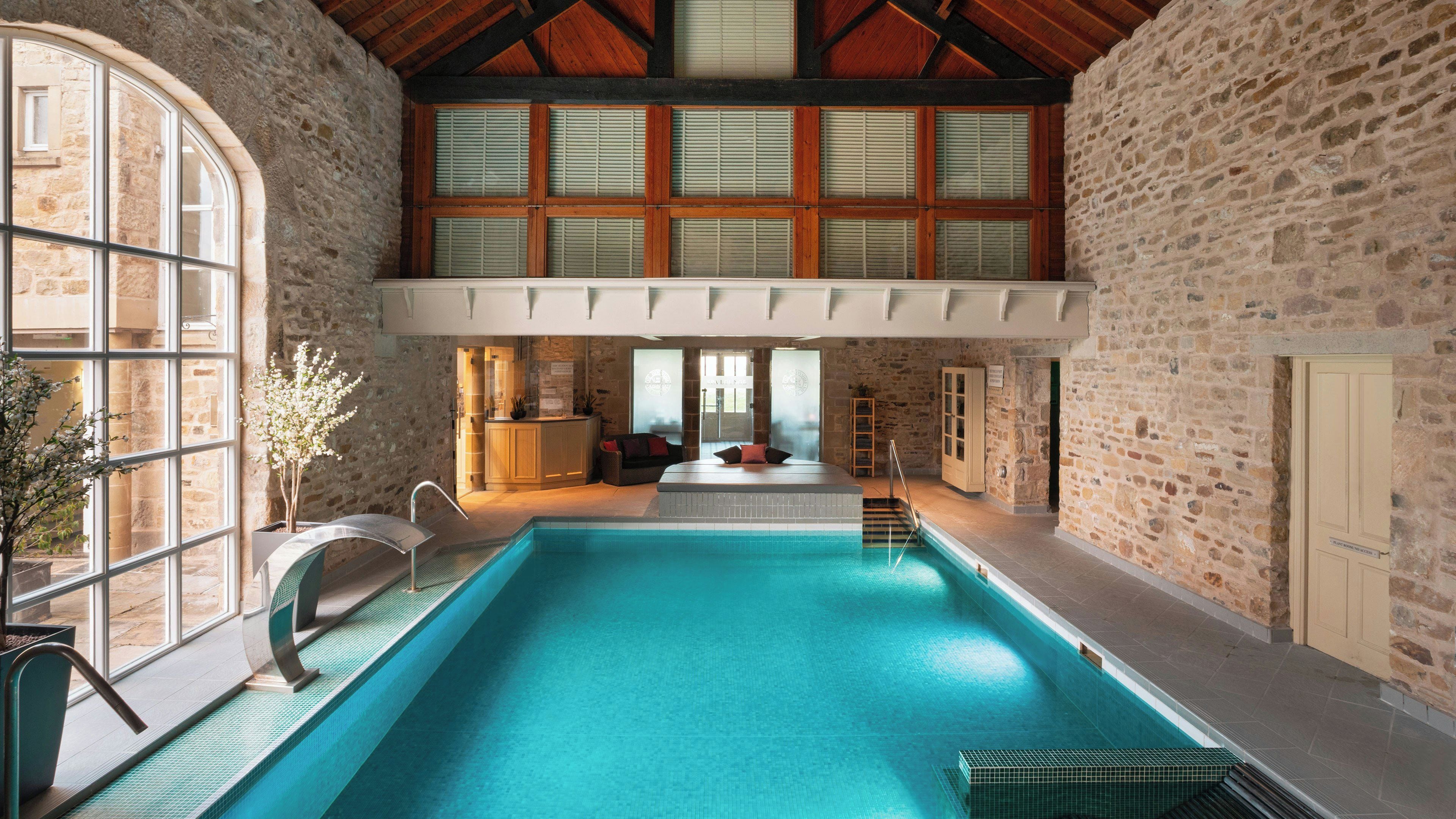 Una amplia piscina interior con pared de piedra y techo de madera, un área de descanso con banco y mesa, y vistas al exterior a través de grandes ventanales.