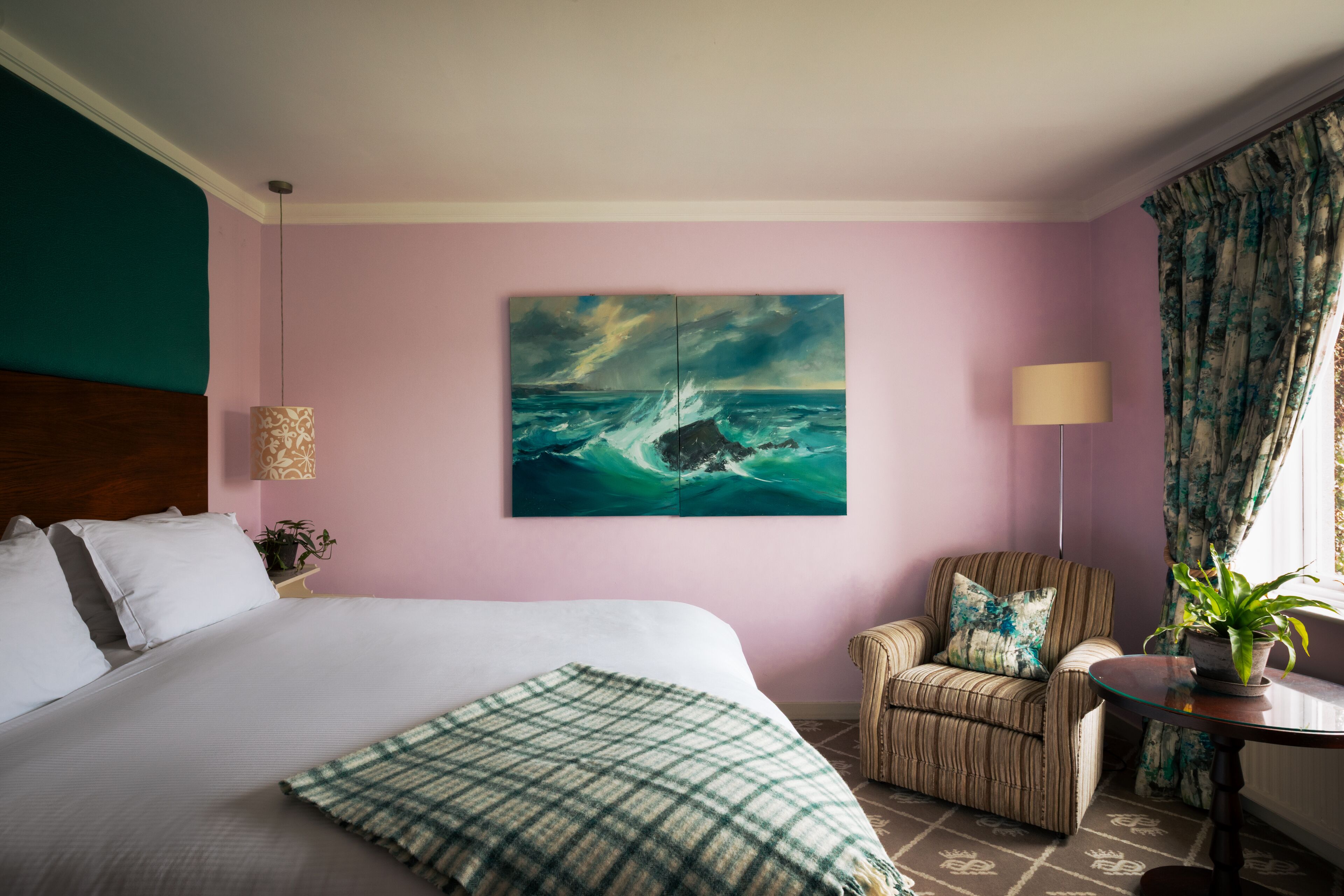 A hotel room with a large bed, a chair, a table with a plant, and a painting of a seascape on the wall.