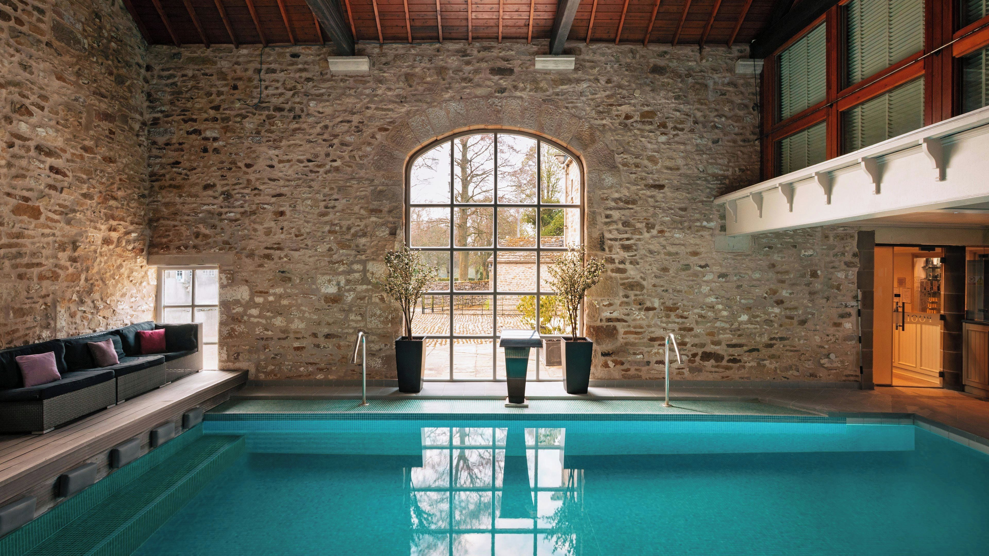 A swimming pool with stone walls and a wooden ceiling, a bench with cushions, and a large arched window.