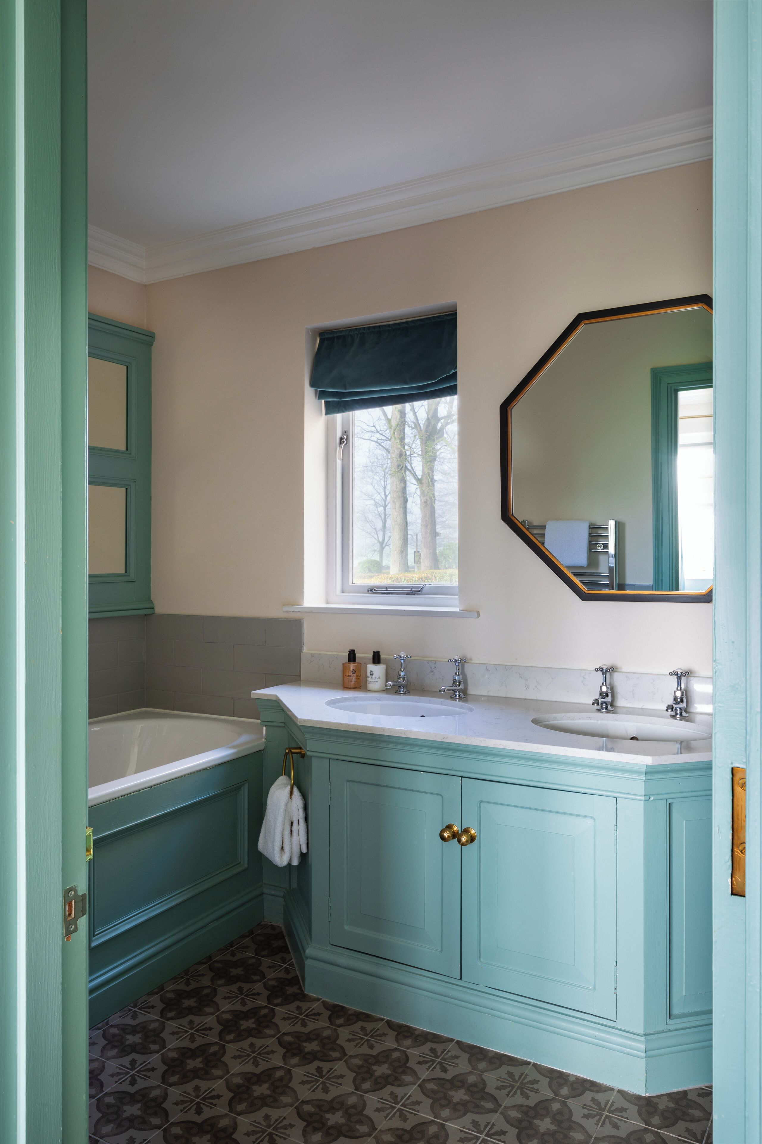 A bathroom with teal cabinetry, a white bathtub, and a window with a view of trees.