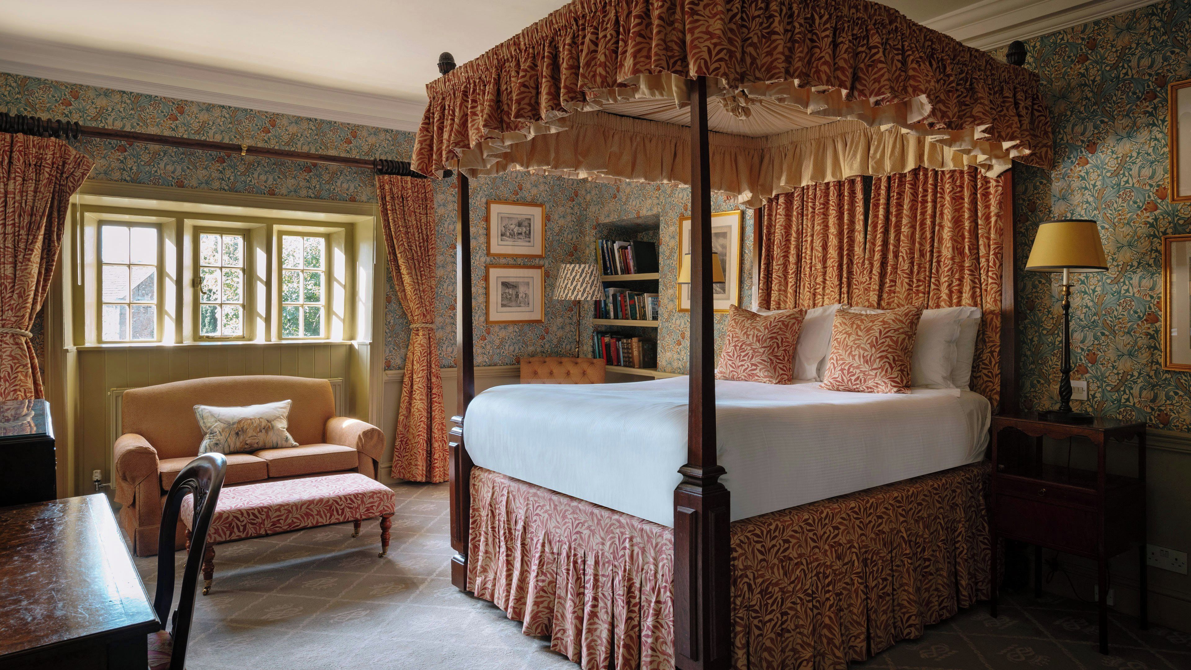 A four-poster canopy bed with a matching canopy, a chaise lounge, a bedside table, a lamp, and a bookshelf in a room with patterned wallpaper and a window.