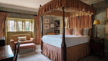 A four-poster canopy bed with a matching canopy, a chaise lounge, a bedside table, a lamp, and a bookshelf in a room with patterned wallpaper and a window.