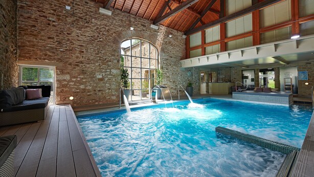 An indoor swimming pool with a stone wall, wooden ceiling, and a seating area with a sofa and cushions. - The Devonshire Arms Hotel (Skipton)