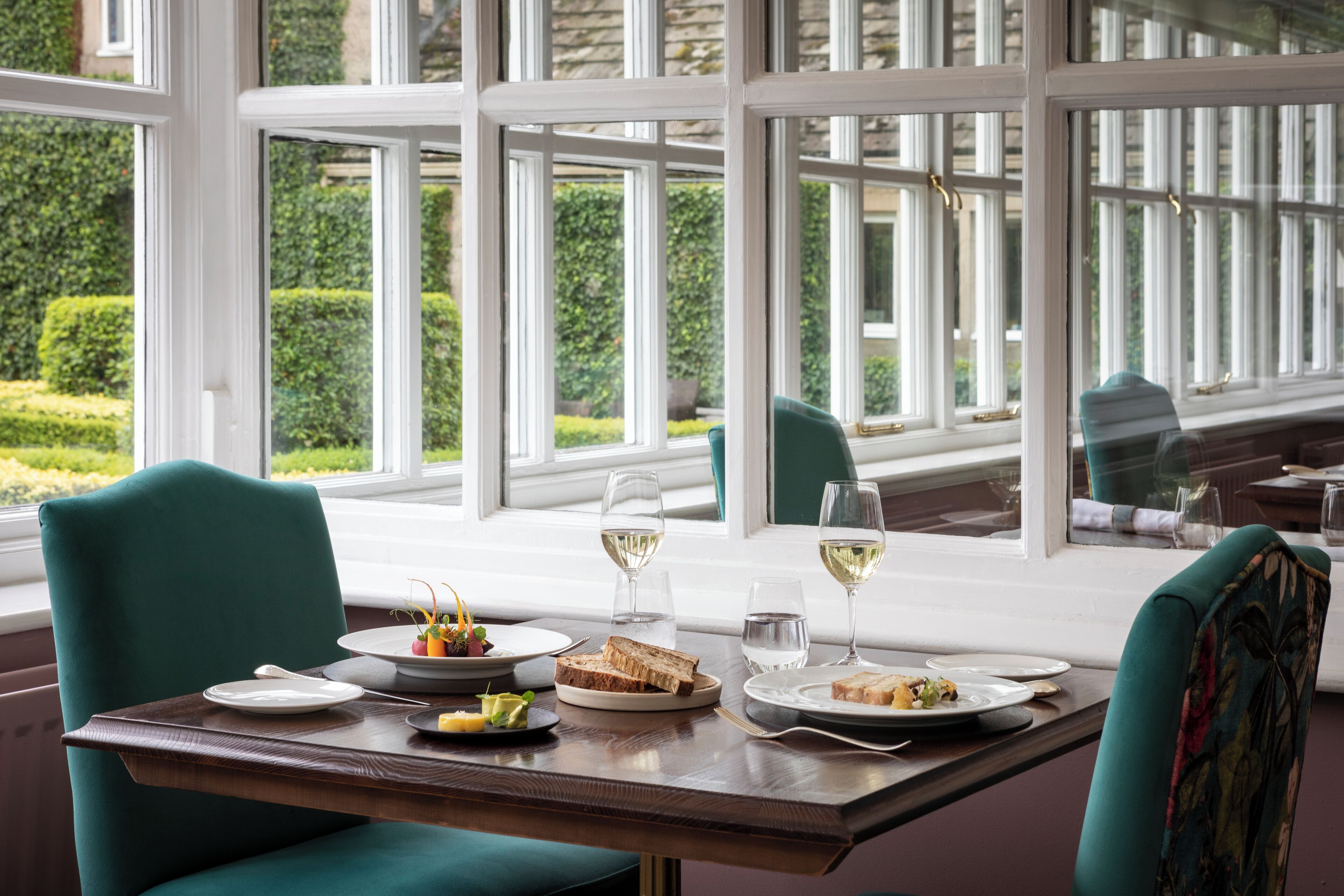 A dining table set with plates, glasses, and cutlery, with a view of greenery outside through large windows.