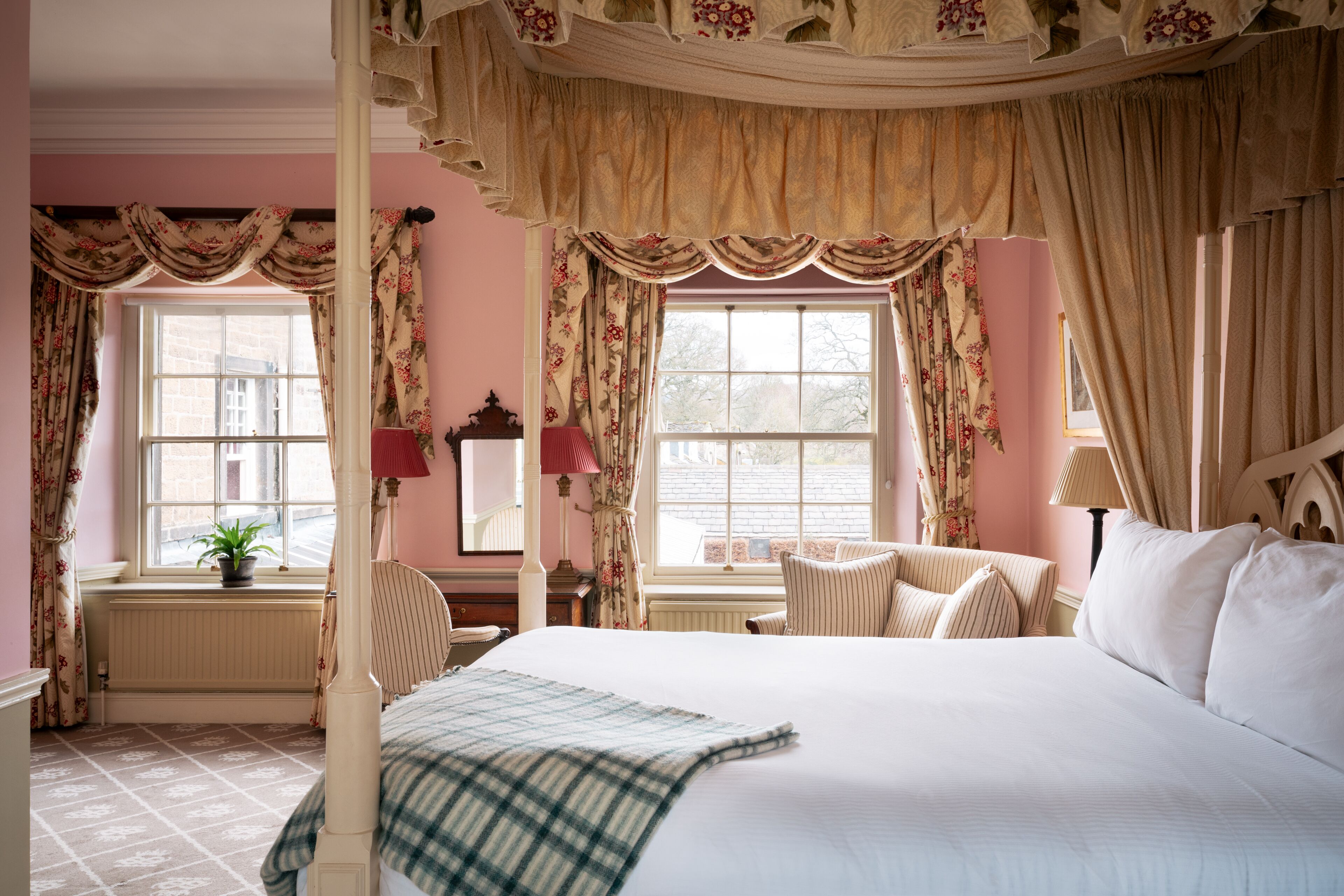 A four-poster bed with a canopy, a large window with floral curtains, a bedside table with a lamp, and a chair with striped cushions.