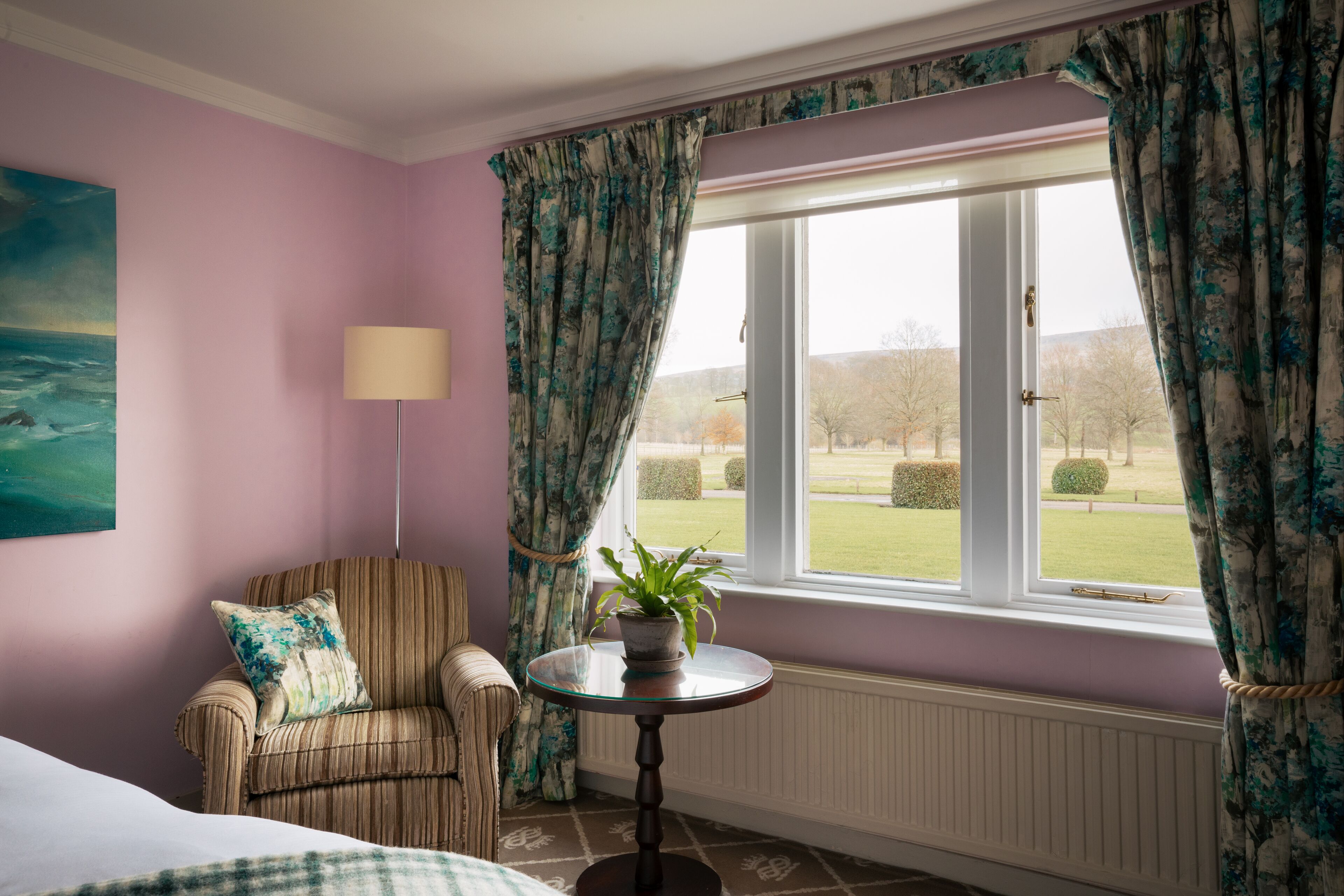 A room with a bed, a chair, a table with a plant, and a window with curtains.
