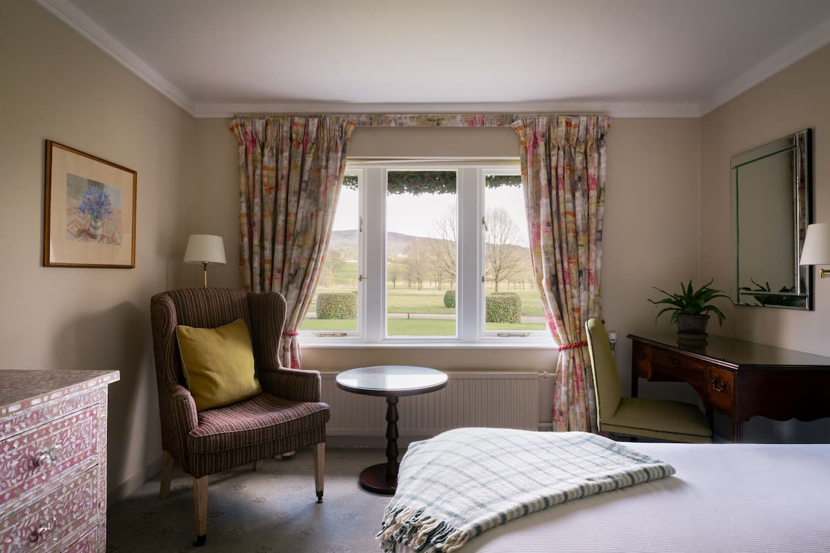 A hotel room with a bed, a chair, a small table, a dresser, a window with curtains, and a framed picture on the wall.