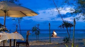 A beach setting with a table set for dining under a large umbrella, surrounded by palm trees and the ocean in the background. - Le Grandeur Balikpapan (Balikpapan)