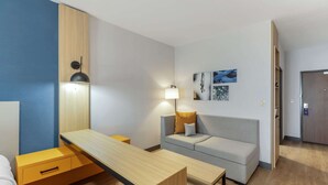 Una habitación de hotel moderna con un escritorio de madera, un sofá gris y una mesita pequeña. Hay una lámpara en el sofá y otra en el escritorio. La habitación tiene una pared de acento azul y piso de madera.