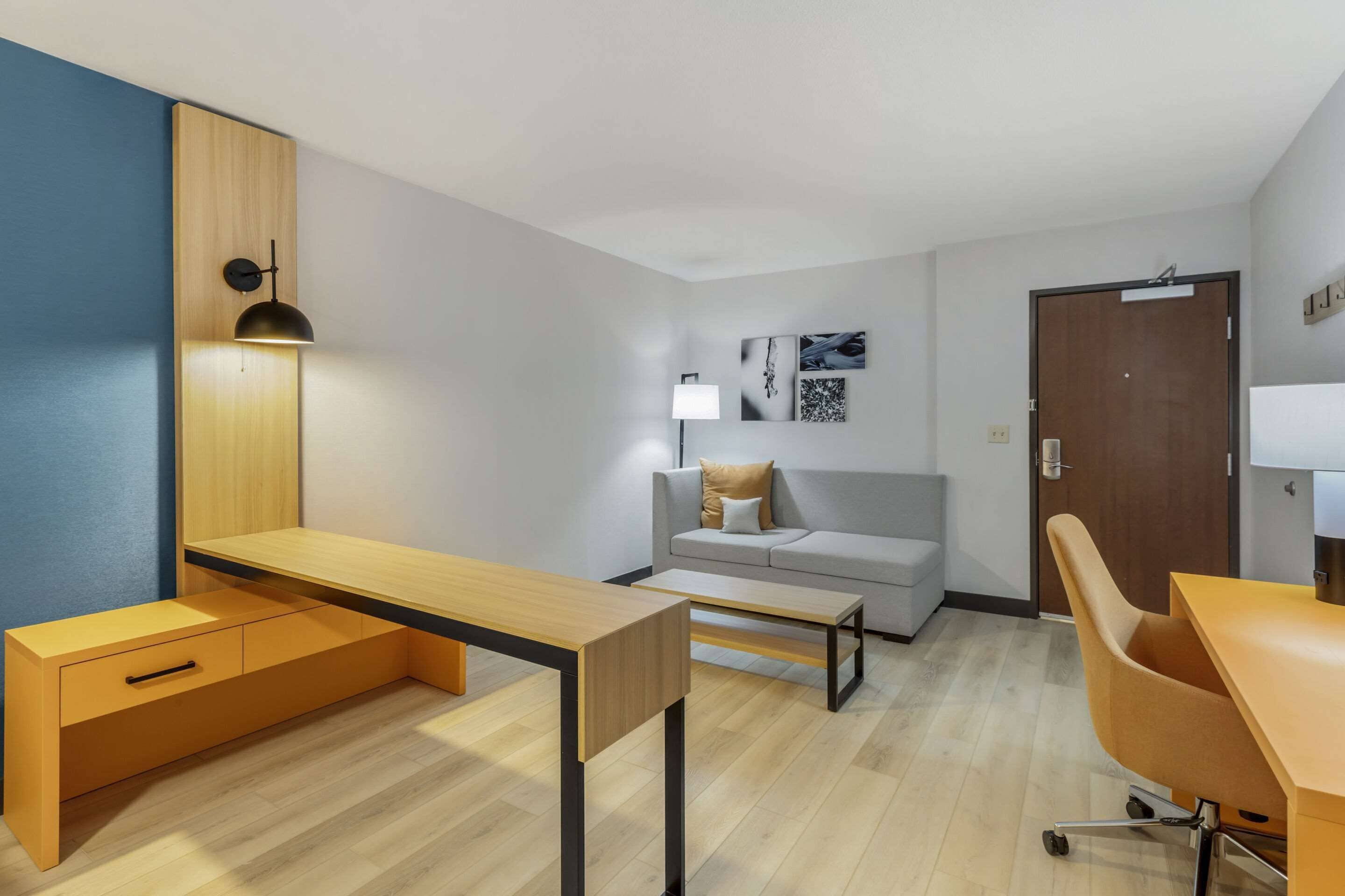 A modern hotel room with a wooden desk, a sofa, a lamp, and a door.