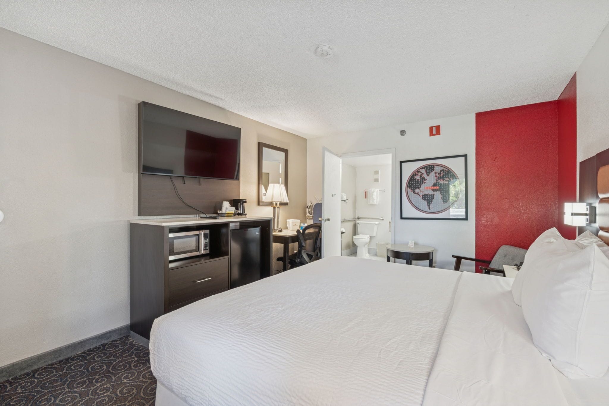 A hotel room with a large bed, a desk with a chair, a television, a microwave, and a small table with a lamp.