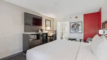 A hotel room with a large bed, a desk with a chair, a television, a microwave, and a small table with a lamp.