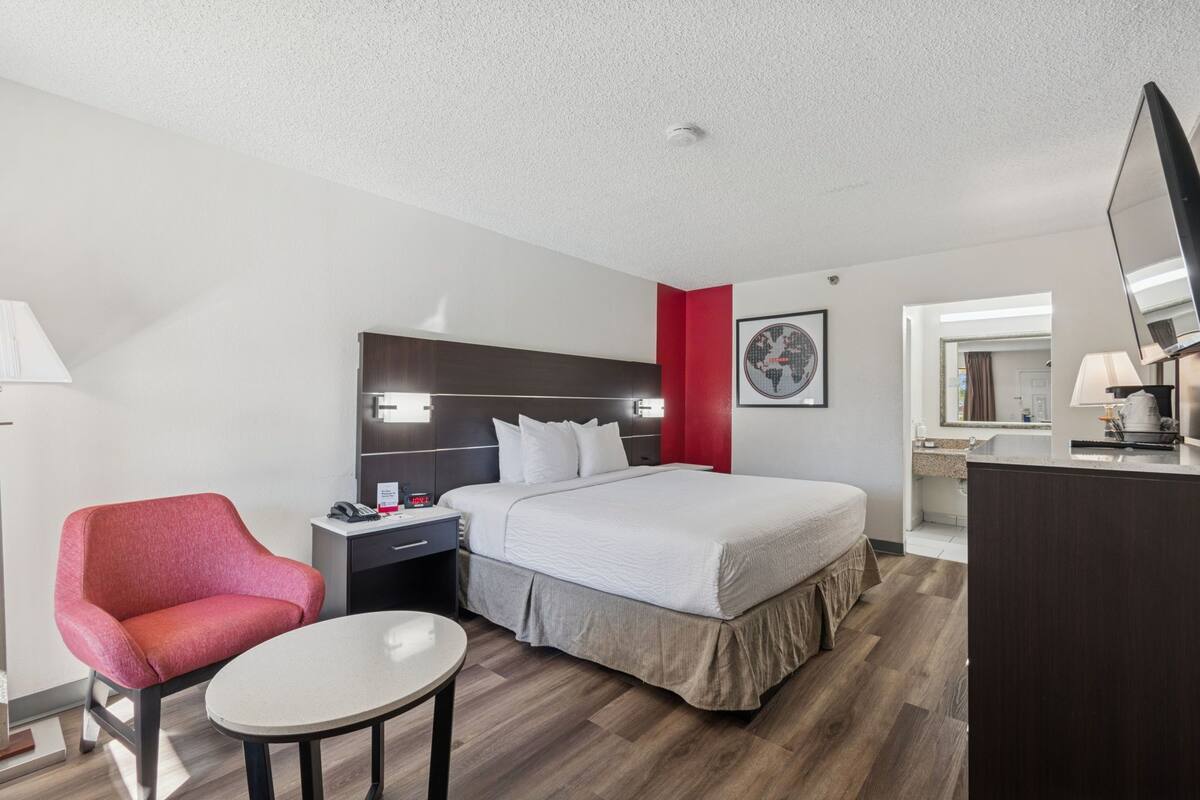 A hotel room with a bed, a desk, a chair, a TV, and a bathroom visible through an open door.