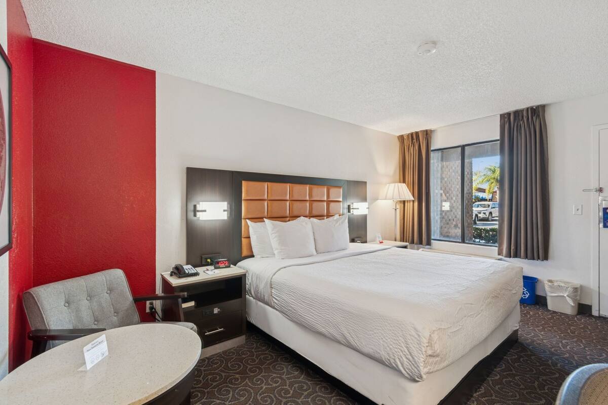 A hotel room with a large bed, a desk with a chair, a small table, a nightstand, and a window with curtains.