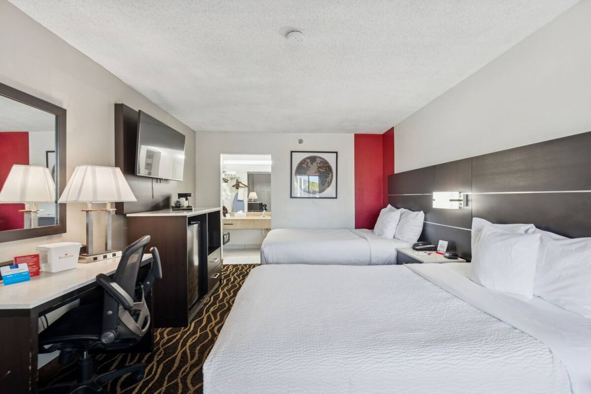 A hotel room with two beds, a desk with a chair, a TV, and a bathroom visible in the background.