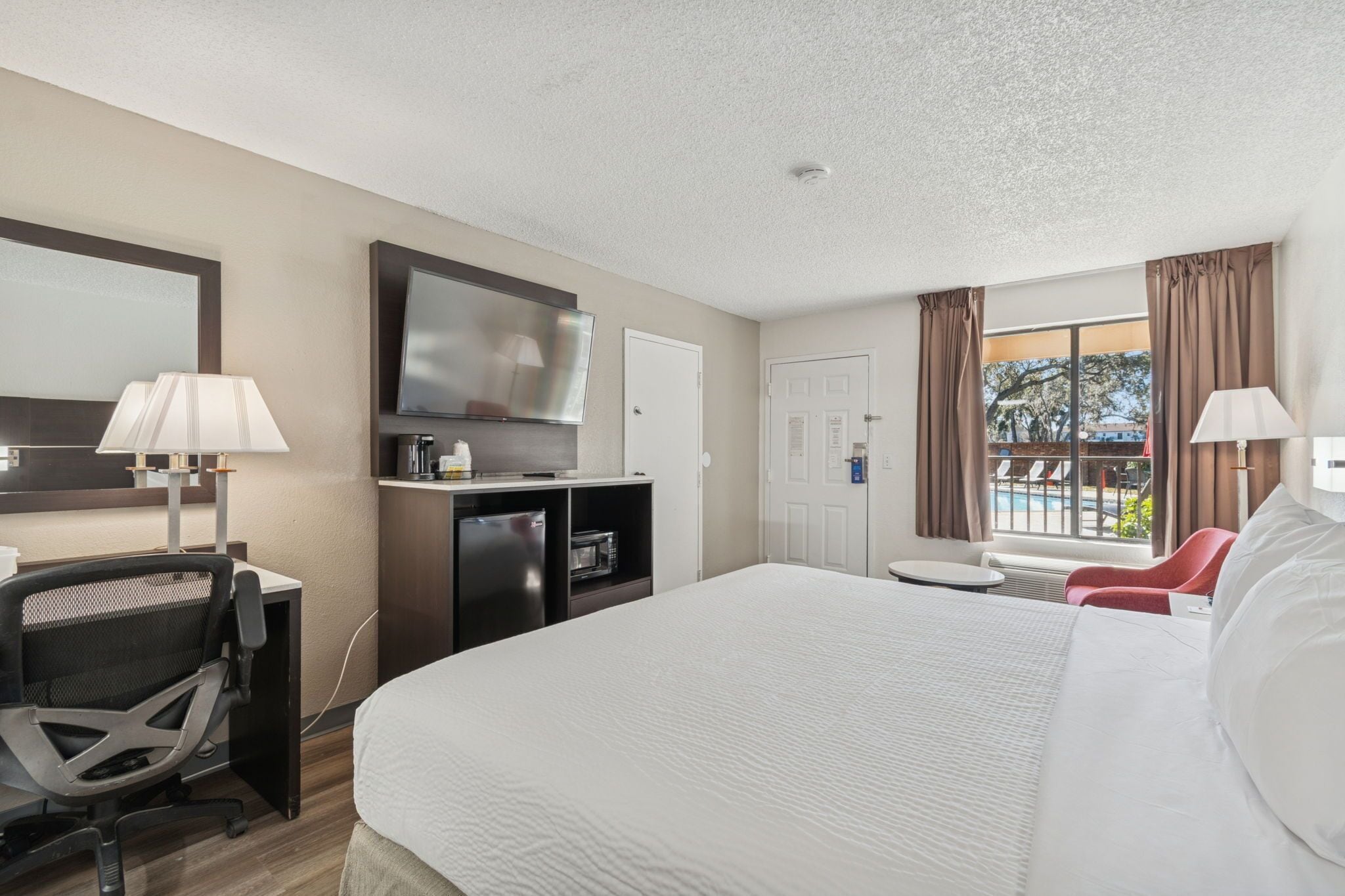 A hotel room with a large bed, a desk with a chair, a television, a minibar, and a window with curtains.